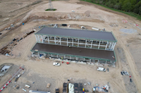 An aerial view of a construction site. A metal framed building with a black roof is prominent.