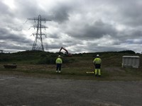 Two contractors in PPE stand in front of an excavator on a grassy slope. Metal transmission towers forming an overhead line are visible in the background.