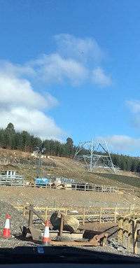 The base of a metal transmission tower under construction, surrounded by rocky slopes and woodland in the distance.