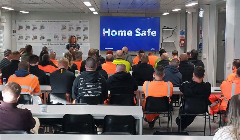 A group of staff in hi vis clothing facing a screen that says "Home Safe"
