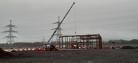A crane next to the metal frame of a building under construction. Several metal transmission towers are visible in the background.
