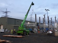 Section of substation infrastructure suspended from a crane. A large green building is present in the background with more substation infrastructure and transmission employees in PPE surrounding it.
