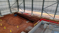 The inside of a metal framed building with a dirt floor, an excavated trench extends from below one of the walls.
