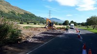 Contractors and an excavator stand by an excavated area next to a surfaced road, filled with rough stones.
