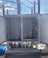 An outdoors metal cabinet with doors open showing large amounts of small electrical equipment fixed inside to the back wall.