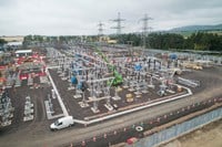 An aerial view of a substation section under construction. A crane and other construction vehicles are present, and transmission infrastructure is visible in the background.