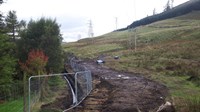 A muddy trench at the base of a grassy slope, various wooden and metal transmission towers are present in the background.
