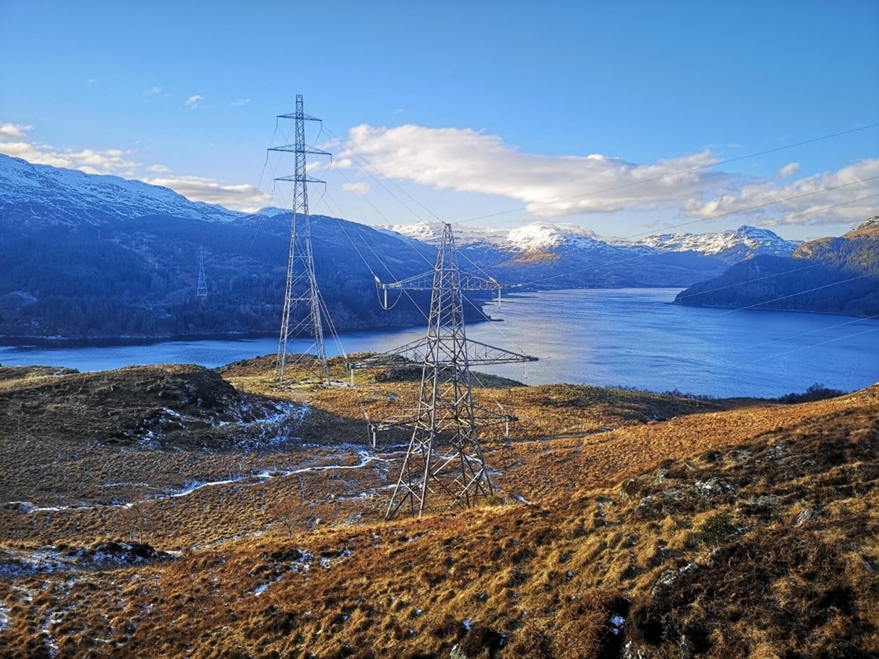 Transmission towers beside a loch with snow-covered mountains in the background