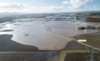 An aerial view of a flooded rural area with transmission towers and an overhead line. There is an annotated yellow line through the area showing a flooded road, titled "Main Access route to rite"