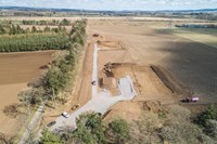 An aerial view of cleared dirt construction area next to an access road. Metal transmission towers forming an overhead line are visible in the background.