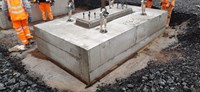 A large concrete cuboid, embedded in a depression in rough construction ground. Large metal fittings extend from the top of the slab, taught straps extend upwards from the fittings out of shot. Several contractors in PPE stand nearby the structure.