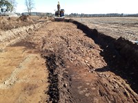 Excavated area of a dirt construction site, an excavator is present at the end of the excavated area.