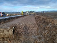 A pipe semi-submerged in dirt and mud in the bottom of an excavated trench next to a road. Gravel is piled against the far side of the pipe and fills the trench stretching away from the foreground. Contractors in PPE drive flattening construction vehicles over the gravel.