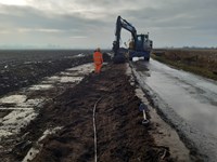 An excavated area next to a road. A contractor in PPE and an excavator are present on the edge of the road.