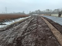 A dirt section of ground between a field and a road. There is a line of light brown gravel alongside the road.