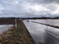 A flooded road, with flooded fields on each side. A car drives down the road with the water up to its' chassis.