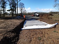 Wire frame and material laid in an excavated dirt road area. Construction vehicles present in the background on a surfaced area.