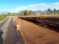 An excavated area next to a road. A construction vehicle is present at the end of the area.