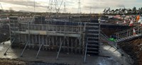 A large excavated area, inside of which metal framed walls are supported by scaffolding. Above and in the background are metal transmission towers forming an overhead line.