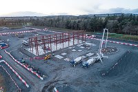 An aerial view of two concrete mixer vehicles near a metal framed building under construction. A crane and several concrete sections of ground surround the mixer vehicles.