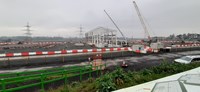 A white metal framed building under construction on a construction site. Cranes and several excavators and construction vehicles surround the building.