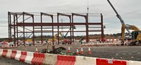 A metal framed building under construction on a construction site.  A crane and several excavators and construction vehicles surround the building.