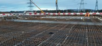 A metal rebar frame within a wide excavated area. Construction vehicles and metal transmission towers are present in the background.