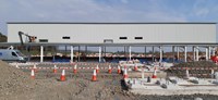 A metal framed building, the top section is clad in corrugated metal. In the foreground sections of concrete are sunk into a dirt area. Construction vehicles can be seen in the background.