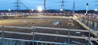 A metal rebar frame within a wide excavated area. Construction vehicles and metal transmission towers are present in the background.