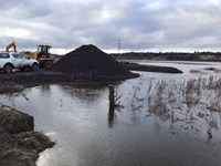 View across a flooded area to a slightly elevated dirt area with a large dirt mound. A construction vehicle and a car are parked there.