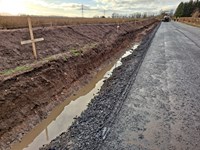 A muddy trench alongside a surfaced road. Contractors in PPE and a construction vehicle are present at the end of the road.