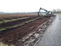 An excavated area next to a road, an excavator is present in the background.