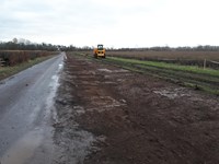 An excavated area next to a road, an excavator is present in the background.