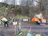 Contractors in woodland, cut logs and a woodchopping machine are nearby.