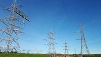 Contractors in PPE on a thin, metal guard-railed platform suspended from a metal transmission tower. Other transmission towers are visible nearby.