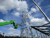 A section of a metal transmission tower suspended from a crane over a transmission tower base. Contractors in PPE are perched on the base.