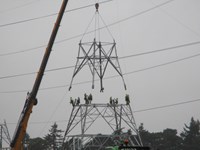 A section of a metal transmission tower suspended from a crane over a transmission tower base. Contractors in PPE are perched on the base.