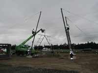 A metal transmission tower under construction using a crane. Several contractors in PPE are present on the tower base.