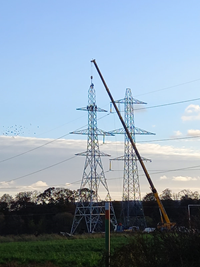 A crane adding the topmost section of a metal transmission tower. Several people in PPE are perched on the top of the tower.