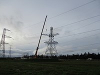 A crane adding a section of a new transmission tower. Several people in PPE are perched on the top of the tower.