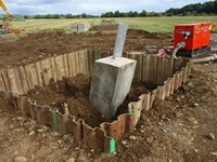 A section of a dirt surfaced ringed by corrugated metal. A concrete block emerges from the dirt and a metal section extends from the concrete.