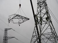 A section of metal transmission tower suspended from a crane at height next to a metal transmission tower. People in PPE stand on the tower.