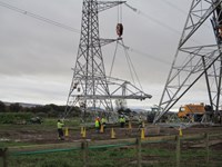 A section of metal transmission tower suspended from a crane. People in PPE stand beneath it. Metal transmission towers under construction are nearby.