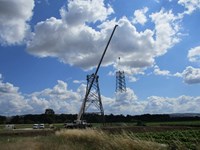 A section of a metal transmission tower suspended from a crane next to a transmission tower under construction. Contractors in PPE are perched atop the tower.