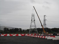 A section of a metal transmission tower suspended from a crane. The section rests on a transmission tower under construction. Contractors in PPE are perched atop the tower.