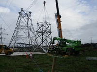 A section of a metal transmission tower suspended from a crane next to a transmission tower under construction. Contractors in PPE are perched atop the tower.