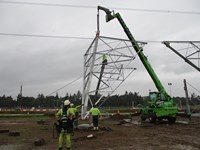 A wedge-shaped corner section of a transmission tower base suspended from a crane. Contractors in PPE on the ground guide the corner to a concrete section on the ground.