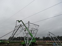 A wedge-shaped corner section of a transmission tower base suspended from a crane. Contractors in PPE on the ground guide the corner to a concrete section on the ground.