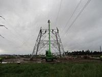 Contractors in PPE atop a base section of a transmission tower under construction. A crane is attached to the top of the section.