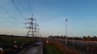 A series of metal transmission towers forming an overhead line. Next to a surfaced road and a fenced construction site.